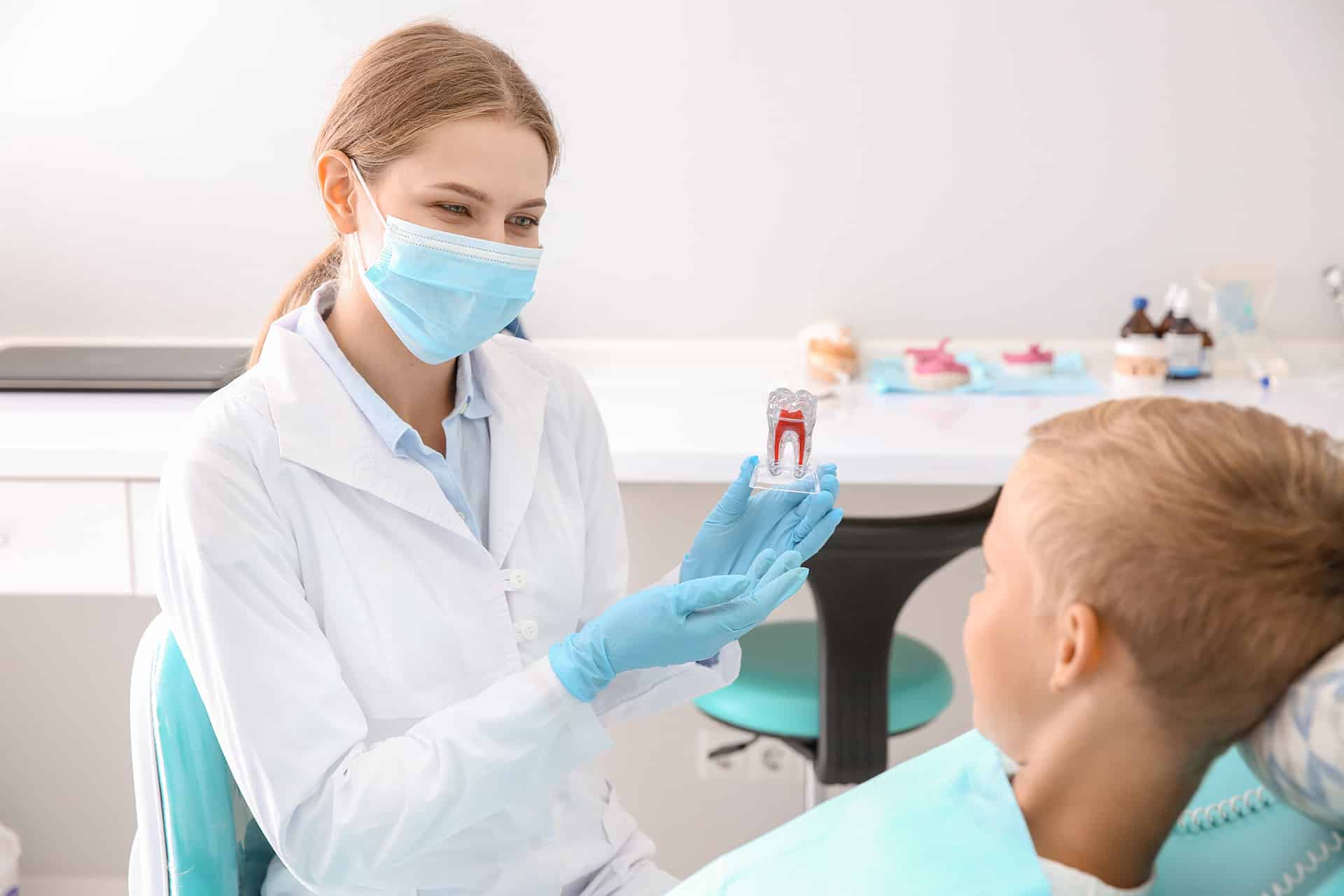 Pannon Kincstár szakmai cikk. Egy hölgy fehér köpenyben és maszkban egy demonstrációs fog modellt mutat egy gyermek páciensnek.