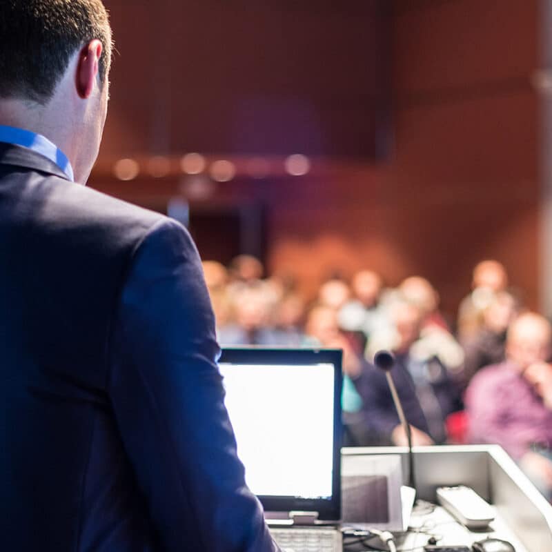 Férfi hátulról látható, ahogyan egy kulturális rendezvényen a számítógépe előtt áll, és beszédet mond. Kulturális rendezvényszervező képzés, Pannon Kincstár szakmai cikk