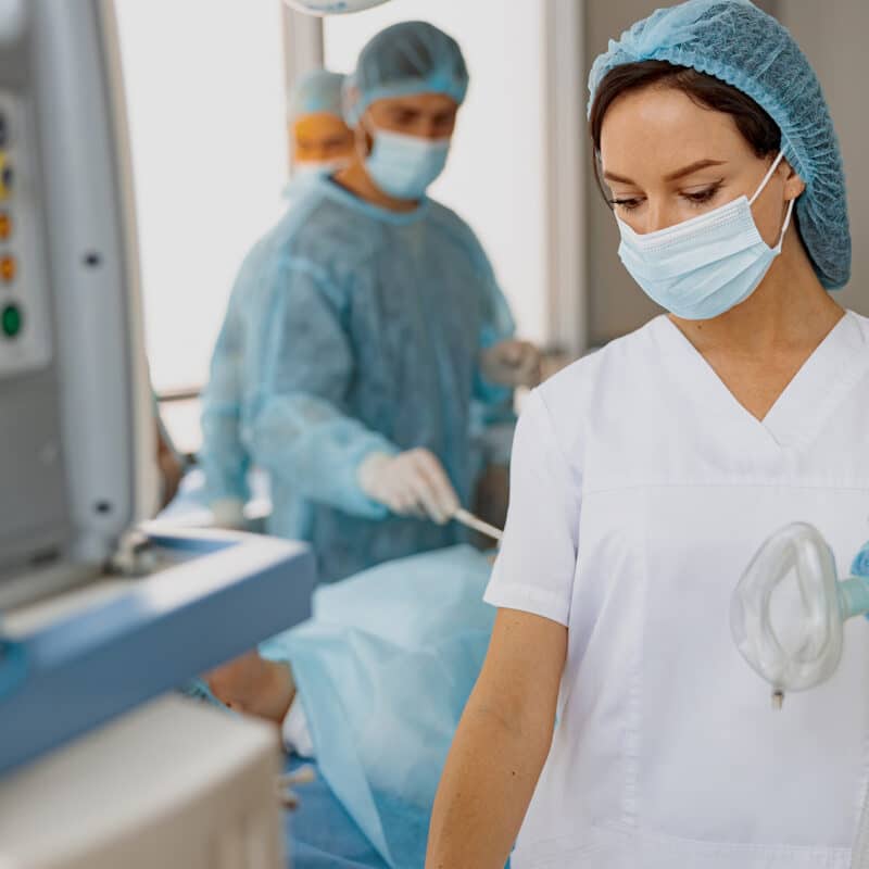 Fiatal nő egészséhügyi ruházatban, maszkban, orvosi műszert a kezében tartva dolgozik. Aneszteziológiai szakasszisztens képzés, Pannon Kincstár szakmai cikk